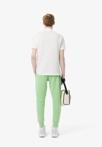Hvid polo skjorte med korte ærmer, grønne sweatpants og en beige taske med sort rem. Modellen har hvide sneakers på. Simpelt design.