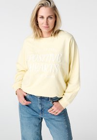 Lichtgele sweatshirt met lange mouwen, bedrukt met witte tekst 'POSITIVE HEARTS' en 'RUE DE PARIS'. Draagt blauwe high-waisted spijkerbroek.