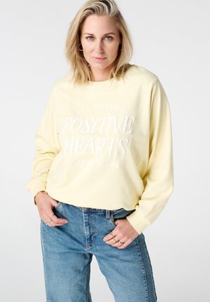 Lichtgele sweatshirt met lange mouwen, bedrukt met witte tekst 'POSITIVE HEARTS' en 'RUE DE PARIS'. Draagt blauwe high-waisted spijkerbroek.