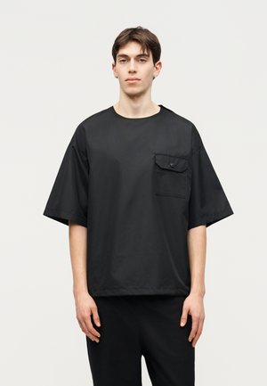 Fiatal férfi laza fekete rövid ujjú ingben, gombos mellzsebbel, áll egy egyszerű világos háttér előtt.