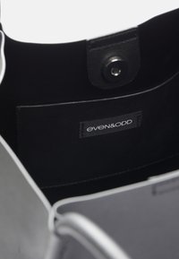 Indvendig i en sort lædertaske med en glat tekstur, en trykknaplukning og et mærke med "even&odd" i hvid tekst.