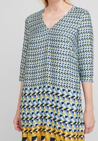 Top con escote en V y patrones geométricos en azul, amarillo y marino, con mangas de tres cuartos y un corte fluido en toda la prenda.