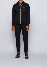 Schwarzer sportlicher Trainingsanzug mit einer Reißverschlussjacke mit goldenen Akzenten, schmal geschnittenen Hosen und schwarzen Turnschuhen mit goldenen Details.