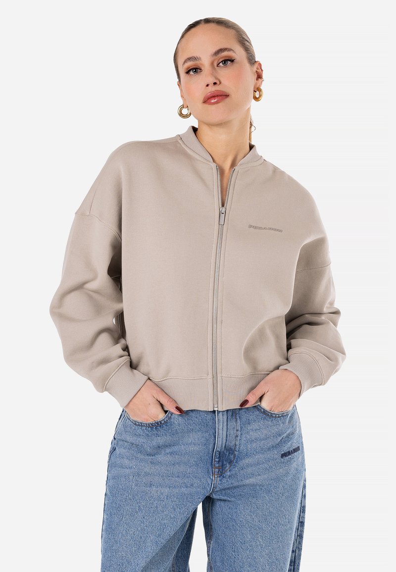 Bežinė zip-up mikrošalio striukė su aukštu apykakle, raukšlėtomis rankovių ir apačios detalėmis, su logotipu ant krūtinės, derinama su mėlynais džinsais.
