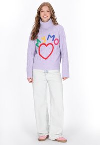 Lila kabelgestrickter Pullover mit hohem Kragen, versehen mit buntem gesticktem Text und Herzdesign. Kombiniert mit hellem Denim und rosa Schuhen.