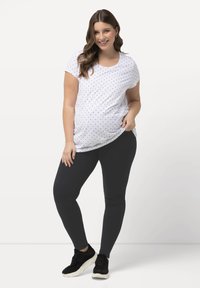 Schwangerschafts-T-Shirt in Weiß mit schwarzen Punkten, aus weichem Stoff, mit kurzen Ärmeln, kombiniert mit schwarzen Leggings und schwarzen Sneakers.