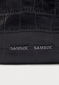 Superficie in pelle nera testurizzata con due loghi metallici "SAMSØE" argento affiancati, evidenziando il dettaglio del marchio sul prodotto.