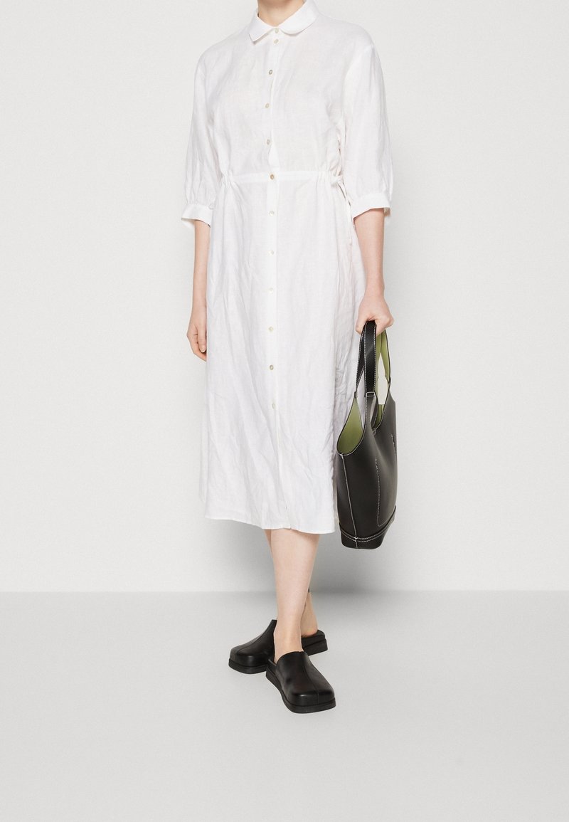 Femme portant une robe-chemise midi blanche boutonnée avec des manches trois-quarts, des chaussures noires sans lacets et tenant un sac fourre-tout noir.