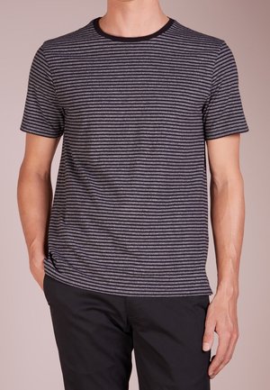 Mężczyzna ubrany w czarno-szary, poziomo prążkowany t-shirt z krótkim rękawem i czarne spodnie, stojący z jedną ręką w kieszeni.