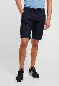 Shorts blu navy con un sottile motivo floreale, tessuto leggero, tasche anteriori e vita con coulisse. Indossati con una maglietta blu e sneakers nere.