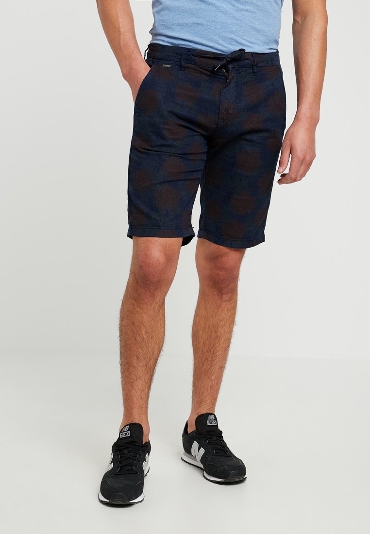 Shorts blu navy con un sottile motivo floreale, tessuto leggero, tasche anteriori e vita con coulisse. Indossati con una maglietta blu e sneakers nere.