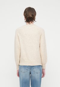 Maglione di lana beige con vestibilità rilassata, polsini e orlo a costine, e maniche raglan, abbinato a jeans azzurri chiari.