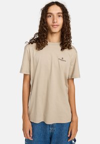Camiseta de algodón beige con cuello redondo y mangas cortas. Presenta un pequeño logo negro en la parte izquierda del pecho. Diseño de corte holgado.