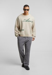 Beige oversized sweater met groene cursieve tekst, gecombineerd met grijze geplooide broek en zwarte schoenen. Model draagt donkere zonnebril.