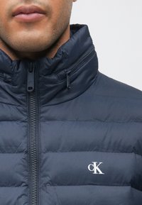 Geacă matlasată albastru închis, cu guler înalt, textură matlasată orizontal, fermoar frontal și logo alb „cK” pe pieptul stâng.