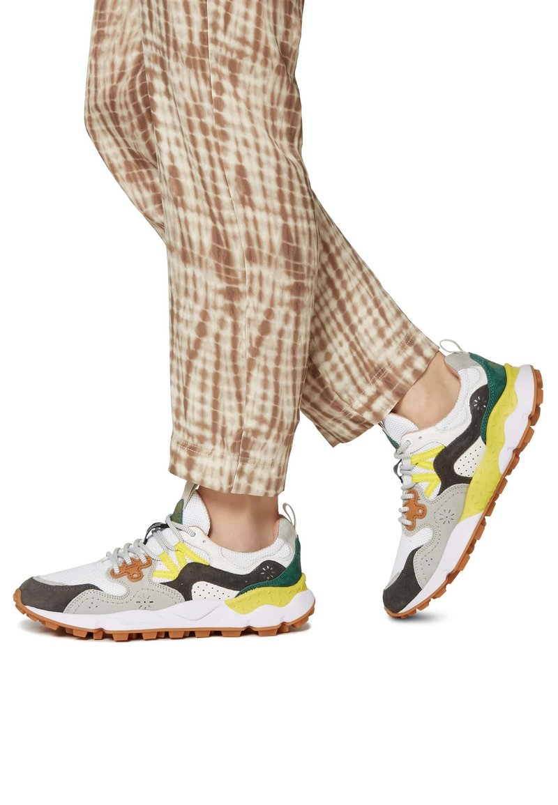 Chaussures de sport multicolores avec une tige en maille blanche, des accents en suède gris et marron, des détails verts et jaunes, et une semelle en caoutchouc texturée.