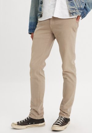 Personne portant un pantalon slim beige, des baskets noires et blanches, une chemise blanche et une veste en jean bleue avec une main dans la poche.