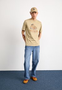 T-shirt en coton beige avec impression graphique, jeans en denim bleu clair et baskets orange avec lacets rouges. Casquette beige avec logo.