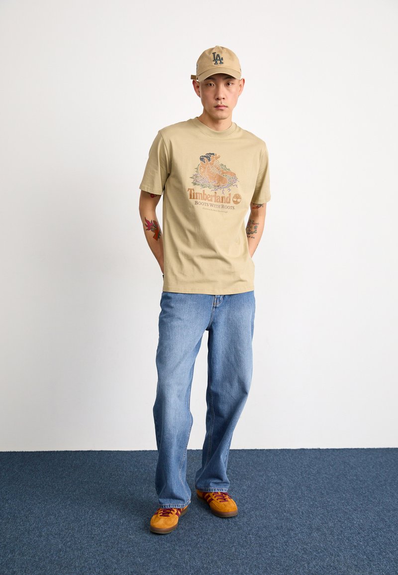 Beige Baumwoll-T-Shirt mit Grafikdruck, hellblaue Jeans und orangefarbene Sneakers mit roten Schnürsenkeln. Beiger Hut mit Logo.