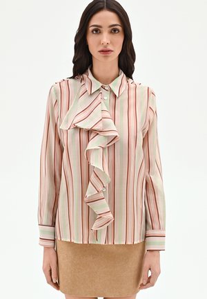 Donna che indossa una camicetta a maniche lunghe con colletto, con righe verticali pastello e dettaglio arricciato sul davanti, abbinata a una gonna color sabbia.