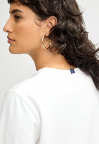 Weißes T-Shirt mit Rundhalsausschnitt und kurzen Ärmeln. Auffälliges marineblaues Etikett hinten. Das Model hat lockige Haare und goldene Creolen.