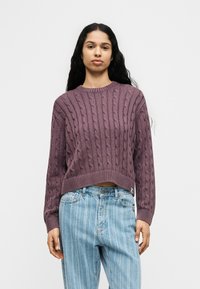 Γυναίκα με μακριά σκούρα μαλλιά που φοράει μοβ πουλόβερ με πλέξη κοτσίδα και ανοιχτό μπλε τζιν με ρίγες και ψηλή μέση, στέκεται μπροστά σε λευκό φόντο.