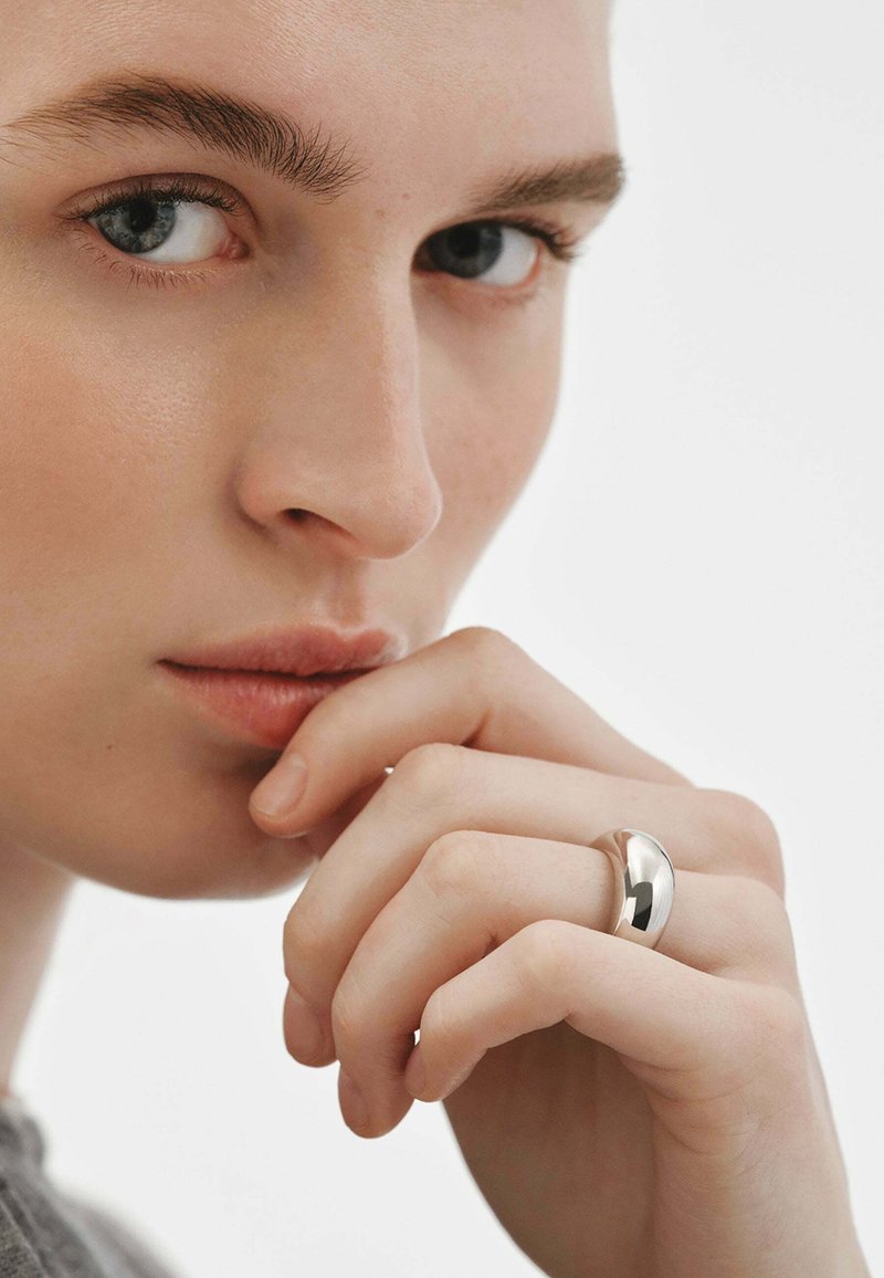 Anillo de plata pulida llevado en una mano, superficie suave con un diseño redondeado. Primer plano de la cara y la mano de una persona, fondo neutro.