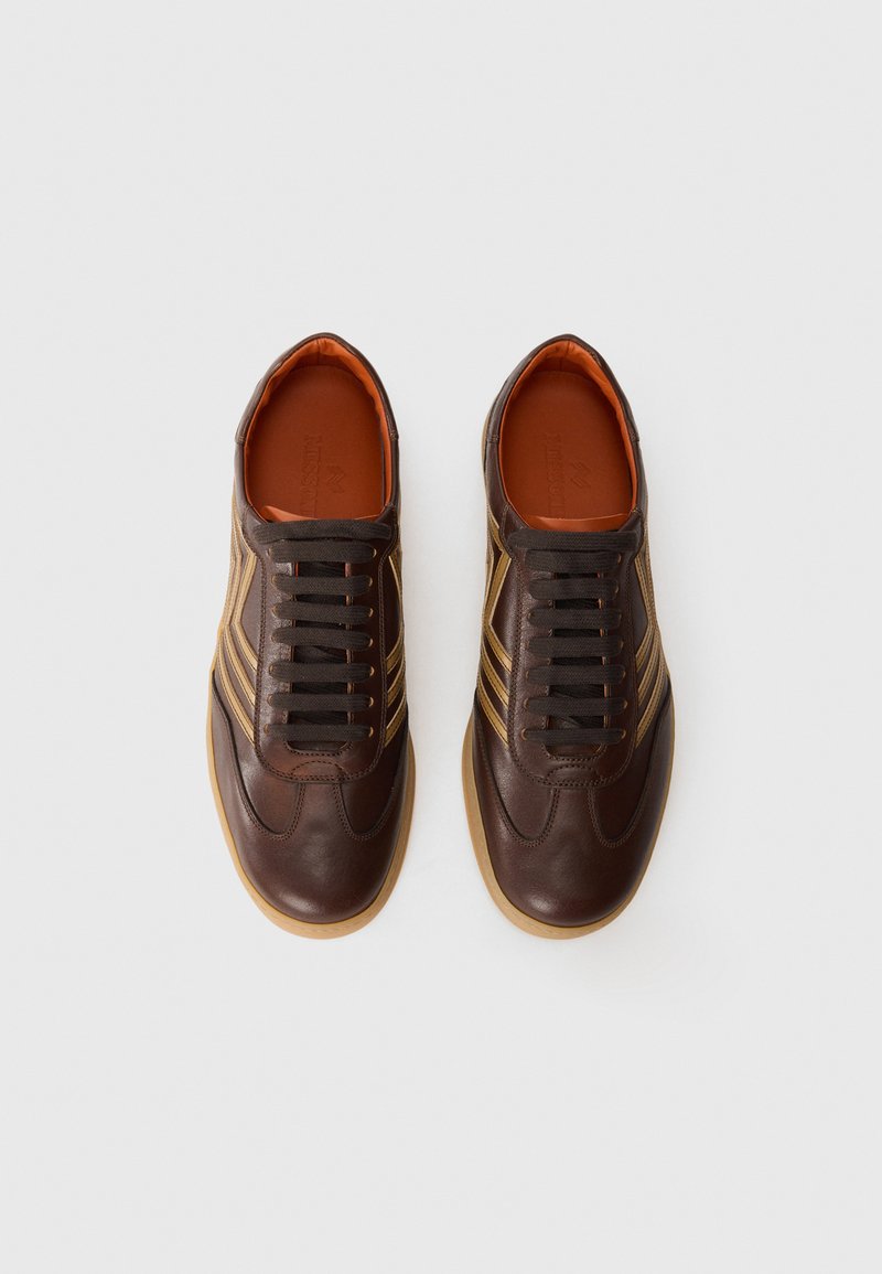 Sneaker in pelle marrone con lacci neri, dotate di tre strisce dorate sui lati e una suola in gomma. L'interno presenta una fodera arancione.
