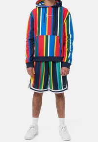 Kolorowa bluza z kapturem w czerwone, niebieskie, zielone i żółte paski, z kieszenią z przodu. W komplecie dopasowane krótkie spodenki w paski i białe trampki.