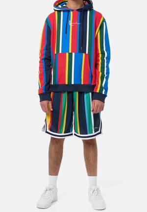 Kolorowa bluza z kapturem w czerwone, niebieskie, zielone i żółte paski, z kieszenią z przodu. W komplecie dopasowane krótkie spodenki w paski i białe trampki.