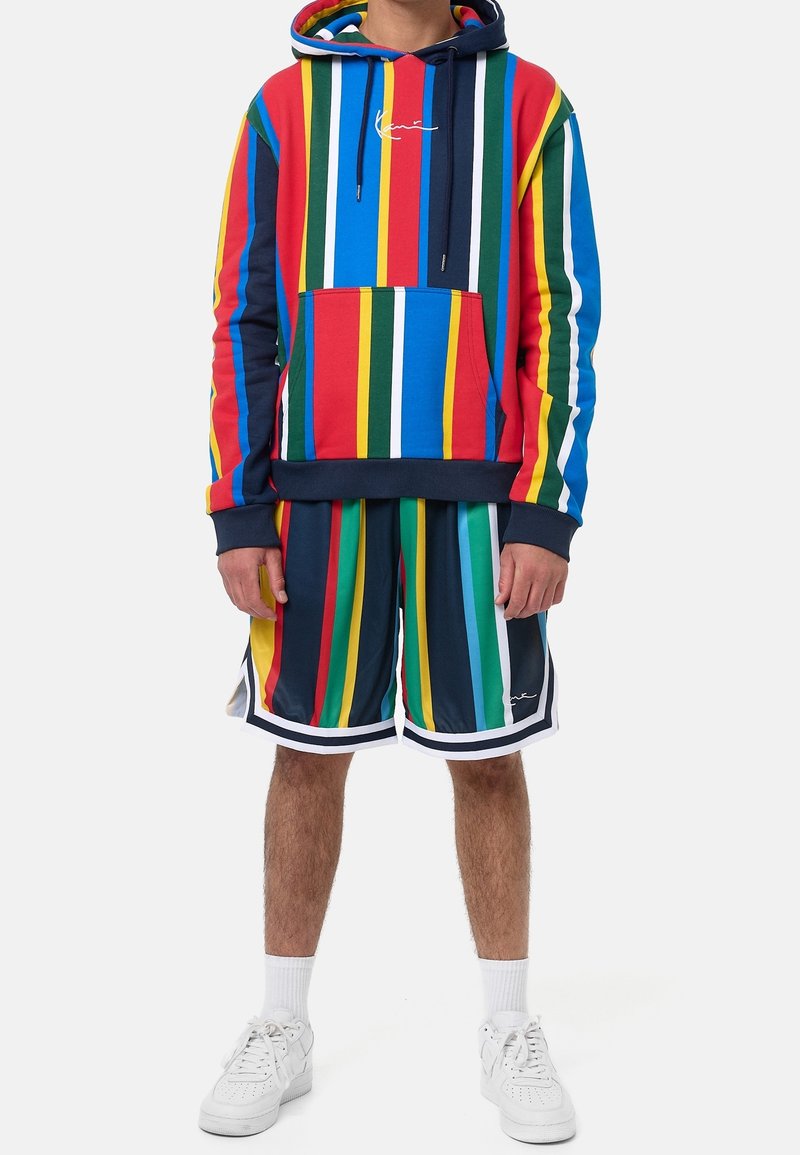 Kolorowa bluza z kapturem w czerwone, niebieskie, zielone i żółte paski, z kieszenią z przodu. W komplecie dopasowane krótkie spodenki w paski i białe trampki.