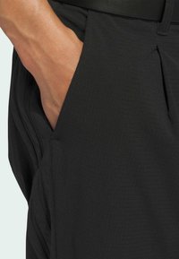 Svarta shorts med texturerad tyg med ett rutmönster och en sidoficka, där en del av en hand vilar i fickan.