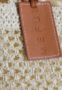Tessuto beige intrecciato con un motivo testurizzato e un'etichetta in pelle marrone con lettere in rilievo. Una cucitura bianca mette in risalto l'etichetta.