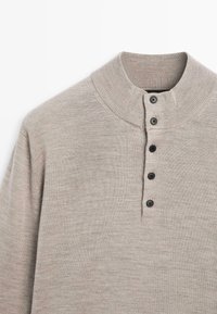 Maglione beige a maglia con colletto a lupetto a coste e quattro bottoni neri allineati verticalmente sul petto.