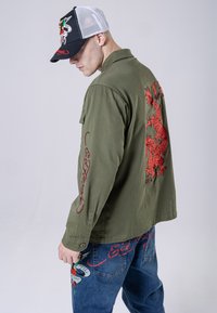 Giacca verde oliva con ricamo di drago rosso sulla schiena e testo rosso sulle maniche, abbinata a pantaloni in denim blu con grafiche colorate.