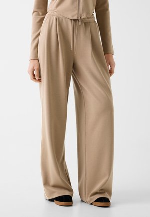 Pantalon beige à jambes larges avec une ceinture élastique et un cordon de serrage, porté avec des chaussures noires à semelles marron sur un fond blanc.