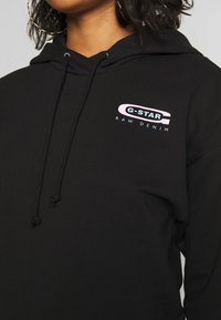 Sudadera con capucha negra que presenta el logo "G-STAR RAW DENIM" en rosa y azul. Fabricada con un tejido suave y con cordones ajustables.
