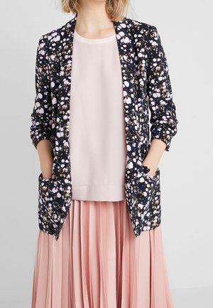 Femme portant un blazer noir avec des taches blanches et marron sur un chemisier rose clair et une jupe plissée rose, les mains dans les poches du blazer.