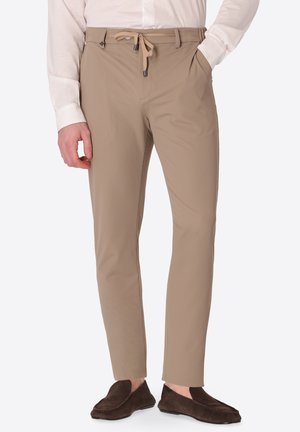 Uomo con pantaloni beige slim-fit con vita con coulisse, camicia bianca e mocassini marrone scuro, in piedi con una mano in tasca.