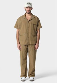 Camisa corta de manga corta marrón con dos bolsillos frontales, combinada con pantalones a juego. Gorra blanca con gráfico de montaña y acentos negros.