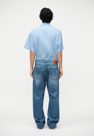 Personne aux cheveux bouclés et foncés, vue de dos, portant une chemise à manches courtes rayée bleu et blanc et un jean bleu ample.