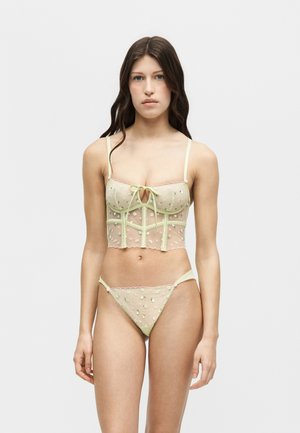 Młoda kobieta ubrana w prześwitujący, jasnozielony zestaw bielizny z kwiatowym haftem, krótki top typu bustier i dopasowane stringi, stojąca na białym tle.