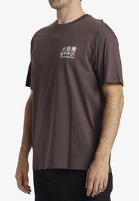 Camiseta de manga corta marrón hecha de algodón, con un diseño gráfico de formas y colores en la parte frontal, etiquetada "BILLABONG."