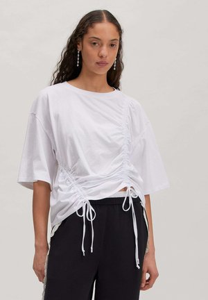 Frau mit langen lockigen Haaren trägt ein weißes, gerafftes Kurzarmtop und schwarze Hose mit Spitzenbesatz an der Seite, stehend vor einem einfarbigen Hintergrund.