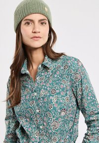 Türkise Bluse mit floralem Muster und Knopfdesign, verziert mit komplexen schwarzen und rosa Mustern. Kombiniert mit einer hellgrünen gerippten Mütze.