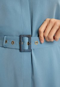 Niebieska sukienka z paskiem, wyróżniająca się gładką teksturą materiału, prostokątną klamrą i złotymi otworami wzdłuż paska.