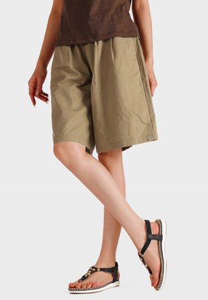 Persona con pantaloncini beige larghi, top marrone e sandali piatti neri con cinturini alla caviglia, in piedi con le gambe incrociate su uno sfondo semplice.