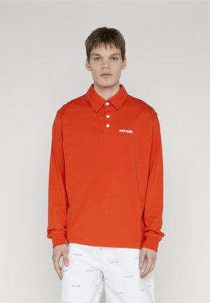 Młody mężczyzna ubrany w jaskrawo pomarańczową, długorękawową koszulkę polo z białym napisem „eckō unltd.” oraz białe spodnie z powtarzającymi się niebieskimi logo.