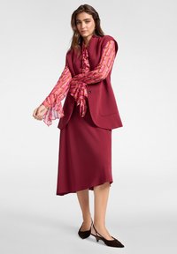 Chaqueta de traje burdeos sobre una blusa rosa con estampado, combinada con una falda a juego. Los detalles incluyen mangas acampanadas y zapatos de tacón negros.