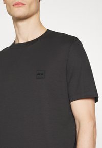 Črna kratka majica s kratkimi rokavi, izdelana iz gladkega materiala, z okroglim izrezom in kvadratnim logotipom na prsih za blagovno znamko.